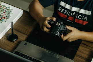 A person is seated at a desk, holding a DSLR camera. On the desk, there is a laptop, a graphic tablet, and a stylus pen. The person wears a dark shirt with the word 'RESILIÊNCIA' printed on it. There is also a piece of artwork on the desk featuring floral designs.