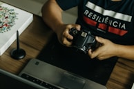 A person is seated at a desk, holding a DSLR camera. On the desk, there is a laptop, a graphic tablet, and a stylus pen. The person wears a dark shirt with the word 'RESILIÊNCIA' printed on it. There is also a piece of artwork on the desk featuring floral designs.