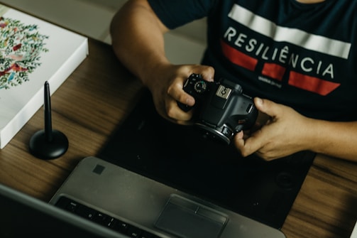 A person is seated at a desk, holding a DSLR camera. On the desk, there is a laptop, a graphic tablet, and a stylus pen. The person wears a dark shirt with the word 'RESILIÊNCIA' printed on it. There is also a piece of artwork on the desk featuring floral designs.