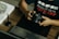 A person is seated at a desk, holding a DSLR camera. On the desk, there is a laptop, a graphic tablet, and a stylus pen. The person wears a dark shirt with the word 'RESILIÊNCIA' printed on it. There is also a piece of artwork on the desk featuring floral designs.