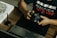 A person is seated at a desk, holding a DSLR camera. On the desk, there is a laptop, a graphic tablet, and a stylus pen. The person wears a dark shirt with the word 'RESILIÊNCIA' printed on it. There is also a piece of artwork on the desk featuring floral designs.