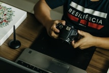 A person is seated at a desk, holding a DSLR camera. On the desk, there is a laptop, a graphic tablet, and a stylus pen. The person wears a dark shirt with the word 'RESILIÊNCIA' printed on it. There is also a piece of artwork on the desk featuring floral designs.