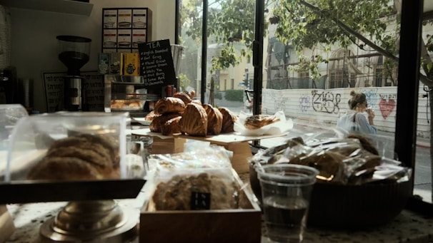 A bakery interior with assorted pastries displayed on a counter near a window. The pastries include croissants and other baked goods in clear packaging. A menu board with tea options is visible in the background, along with a coffee grinder and various decor items. Outside the window, there is a tree and a person sitting at a table, with graffiti on a nearby wall.
