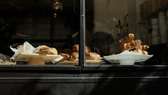 A display window with various pastries, primarily croissants, arranged on plates and stands. The scene appears to be taken from outside the window of a bakery or café. The interior is dimly lit, with other items such as stacked cups and bottles also visible in the background.