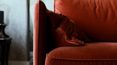 A close-up view of a plush, red velvet sofa with a matching pillow. The background shows part of a side table and a wall with a textured finish.