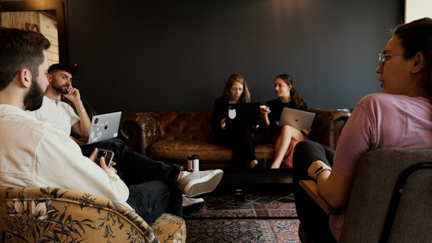 A group of people are seated in a casual office environment. Two individuals are using laptops while others are engaged in conversation. The room features a dark wall, a leather couch, and a patterned rug. The atmosphere suggests a collaborative or meeting setting.