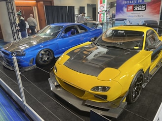 Two modified sports cars are displayed indoors. The car on the left is blue with a carbon fiber hood and custom wheels, while the car on the right is yellow with black accents and a large hood vent. Several people are walking in the background, and promotional banners are visible.