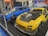 Two modified sports cars are displayed indoors. The car on the left is blue with a carbon fiber hood and custom wheels, while the car on the right is yellow with black accents and a large hood vent. Several people are walking in the background, and promotional banners are visible.