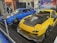 Two modified sports cars are displayed indoors. The car on the left is blue with a carbon fiber hood and custom wheels, while the car on the right is yellow with black accents and a large hood vent. Several people are walking in the background, and promotional banners are visible.