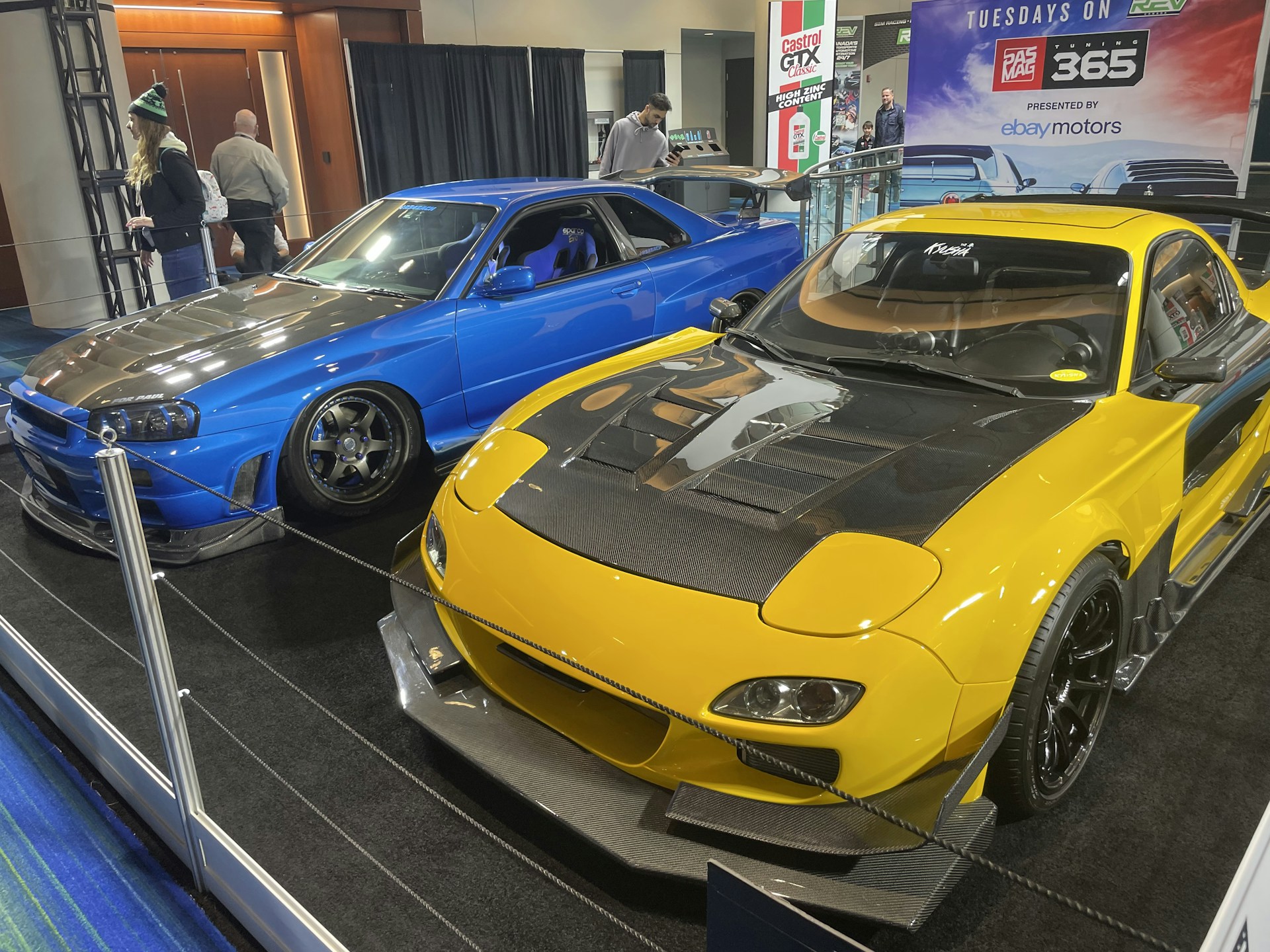 Two modified sports cars are displayed indoors. The car on the left is blue with a carbon fiber hood and custom wheels, while the car on the right is yellow with black accents and a large hood vent. Several people are walking in the background, and promotional banners are visible.