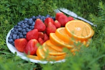 A white plate filled with fresh fruit, including sliced oranges, plump strawberries, and blueberries, is placed on a bed of green grass. The vibrant colors of the fruit contrast beautifully against the lush greenery.
