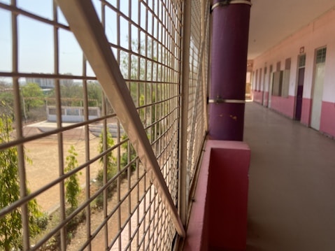 A long corridor with a colorful railing and grid-like metal fencing looking out onto a schoolyard. The corridor has pink and white walls with a uniform design featuring doors aligned in a sequence. Outside, some trees and a sports field can be seen, suggesting an educational facility.