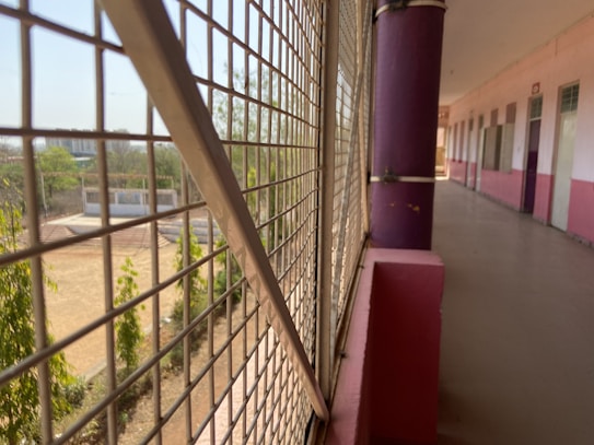 A long corridor with a colorful railing and grid-like metal fencing looking out onto a schoolyard. The corridor has pink and white walls with a uniform design featuring doors aligned in a sequence. Outside, some trees and a sports field can be seen, suggesting an educational facility.