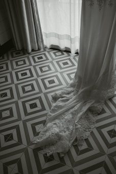 A lace-trimmed white garment, possibly a wedding dress, cascades elegantly onto a geometric patterned floor. Soft light filters through partially opened curtains, creating a serene and delicate atmosphere.