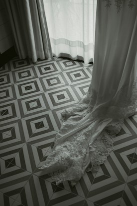 A lace-trimmed white garment, possibly a wedding dress, cascades elegantly onto a geometric patterned floor. Soft light filters through partially opened curtains, creating a serene and delicate atmosphere.
