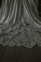 A detailed view of an elegant fabric with intricate floral lace patterns. The fabric drapes gracefully, showcasing its delicate design and texture on a flat wooden surface.