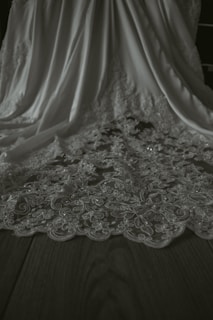 A detailed view of an elegant fabric with intricate floral lace patterns. The fabric drapes gracefully, showcasing its delicate design and texture on a flat wooden surface.