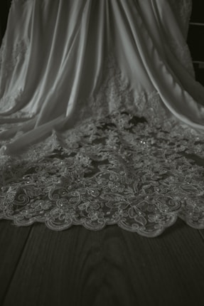 A detailed view of an elegant fabric with intricate floral lace patterns. The fabric drapes gracefully, showcasing its delicate design and texture on a flat wooden surface.