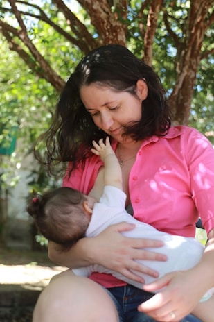A woman in a pink shirt is sitting outdoors under a tree, breastfeeding a baby. Her dark hair is loose, and she looks down affectionately at the infant in her arms. The background consists of lush green foliage and dappled sunlight.
