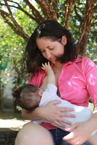 A woman in a pink shirt is sitting outdoors under a tree, breastfeeding a baby. Her dark hair is loose, and she looks down affectionately at the infant in her arms. The background consists of lush green foliage and dappled sunlight.