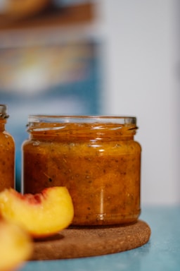 Close-up of a rustic jar filled with golden peach BBQ sauce surrounded by fresh peaches and spices on a wooden table.