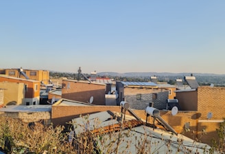 A residential area with numerous brick buildings and satellite dishes on the rooftops. The structures are closely packed with a clear blue sky overhead. Bushes and plant life can be seen in the foreground, while a distant landscape and tree line stretch across the horizon.
