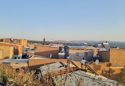 A residential area with numerous brick buildings and satellite dishes on the rooftops. The structures are closely packed with a clear blue sky overhead. Bushes and plant life can be seen in the foreground, while a distant landscape and tree line stretch across the horizon.