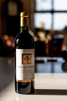 A bottle of Château L'Évêché Pomerol wine from 2019 stands on a smooth surface, illuminated by soft natural light. Its label is elegantly designed with gold accents and intricate typography, indicating its status as a Bordeaux grand vin. The background is softly blurred, suggesting a warm, cozy setting, possibly a dining area or kitchen.