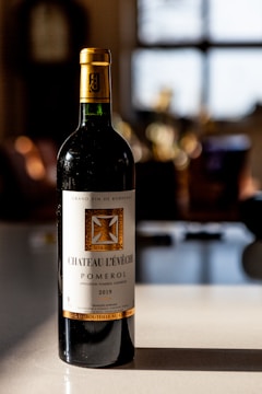 A bottle of Château L'Évêché Pomerol wine from 2019 stands on a smooth surface, illuminated by soft natural light. Its label is elegantly designed with gold accents and intricate typography, indicating its status as a Bordeaux grand vin. The background is softly blurred, suggesting a warm, cozy setting, possibly a dining area or kitchen.
