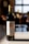 A bottle of Château L'Évêché Pomerol wine from 2019 stands on a smooth surface, illuminated by soft natural light. Its label is elegantly designed with gold accents and intricate typography, indicating its status as a Bordeaux grand vin. The background is softly blurred, suggesting a warm, cozy setting, possibly a dining area or kitchen.