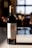 A bottle of Château L'Évêché Pomerol wine from 2019 stands on a smooth surface, illuminated by soft natural light. Its label is elegantly designed with gold accents and intricate typography, indicating its status as a Bordeaux grand vin. The background is softly blurred, suggesting a warm, cozy setting, possibly a dining area or kitchen.