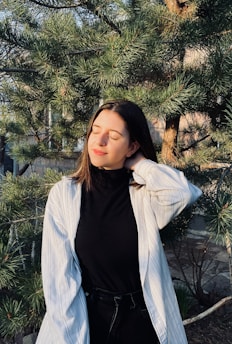 A person with long hair, wearing a black top and a light-colored striped shirt, stands with their eyes closed and a serene expression. They are surrounded by lush green pine branches, with sunlight creating a warm glow on their face.