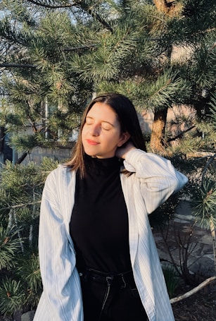 A person with long hair, wearing a black top and a light-colored striped shirt, stands with their eyes closed and a serene expression. They are surrounded by lush green pine branches, with sunlight creating a warm glow on their face.