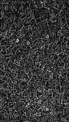 A densely packed arrangement of numerous black screws with a sharp, pointed end and a flat, cross-shaped head. The screws are piled together in a way that their heads and threads interlock, creating a textured, industrial aesthetic.