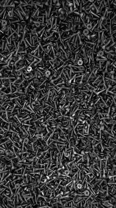 A densely packed arrangement of numerous black screws with a sharp, pointed end and a flat, cross-shaped head. The screws are piled together in a way that their heads and threads interlock, creating a textured, industrial aesthetic.
