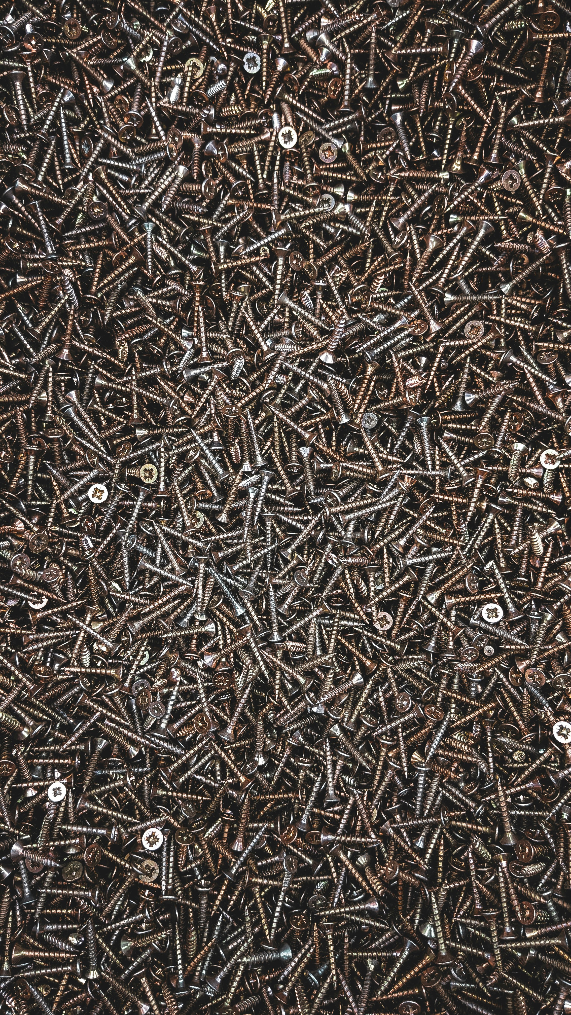 A densely packed assemblage of metal screws, overlapping and haphazardly arranged. The screws are dark in color, with a glossy appearance, and vary slightly in size and type. Some screws have rounded heads while others are flat.
