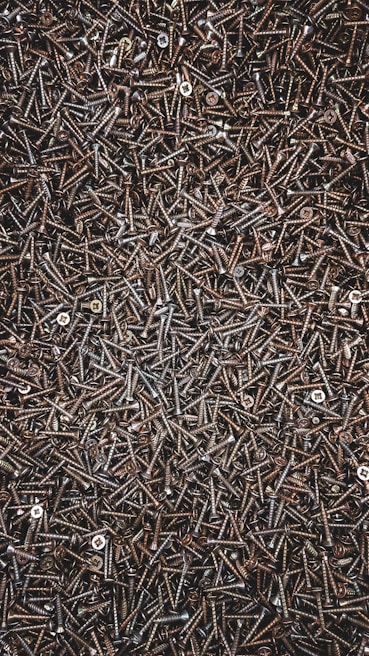 A densely packed assemblage of metal screws, overlapping and haphazardly arranged. The screws are dark in color, with a glossy appearance, and vary slightly in size and type. Some screws have rounded heads while others are flat.