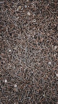 A densely packed assemblage of metal screws, overlapping and haphazardly arranged. The screws are dark in color, with a glossy appearance, and vary slightly in size and type. Some screws have rounded heads while others are flat.