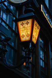 An intricate, vintage-style lamp hangs from a building facade. The lamp's design features colored glass panels with decorative red and yellow patterns, emitting a warm glow. The surrounding building includes dark wooden frames and reflects a dim urban setting.