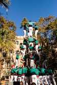 A human tower is being formed outdoors with people dressed in green shirts and white pants. Several tiers of individuals are standing on each other's shoulders to create a tall, multi-level structure. The scene is set against a backdrop of trees and a building under a clear blue sky.