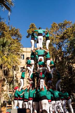 A human tower is being formed outdoors with people dressed in green shirts and white pants. Several tiers of individuals are standing on each other's shoulders to create a tall, multi-level structure. The scene is set against a backdrop of trees and a building under a clear blue sky.