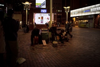 A street performance scene at night with three musicians sitting on the pavement, playing musical instruments. The area is dimly lit, creating a cozy atmosphere, while a few people watch or pass by. The background features a lighted sign and entrance to a commercial area.