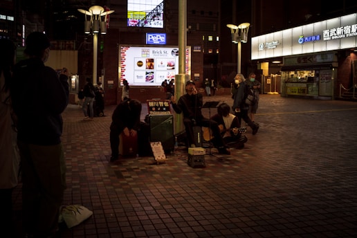 A street performance scene at night with three musicians sitting on the pavement, playing musical instruments. The area is dimly lit, creating a cozy atmosphere, while a few people watch or pass by. The background features a lighted sign and entrance to a commercial area.