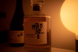 A bottle of Hibiki Suntory Whisky is prominently featured against a warm, dimly lit background. To the left, there is another bottle, partially visible, and on the right, a soft glowing light creates an intimate atmosphere.