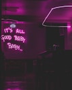 A dimly lit room with a neon sign that reads 'IT'S ALL GOOD BABY BABY..' in bright pink. There is a minimalist interior design featuring a bar counter, chairs, and a modern ceiling light fixture.