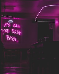 A dimly lit room with a neon sign that reads 'IT'S ALL GOOD BABY BABY..' in bright pink. There is a minimalist interior design featuring a bar counter, chairs, and a modern ceiling light fixture.
