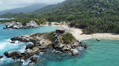 A lush coastal landscape with a rocky outcrop extending into turquoise waters. Atop the rocks is a thatched-roof structure surrounded by visitors. A sandy beach lined with palm trees is visible to the right, while dense green hills rise in the background.