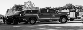 A black and white image featuring multiple emergency response vehicles, including a fire truck marked 'Waco Fire Battalion Chief' and another vehicle labeled 'Elm Mott'. The scene is outdoors with trees in the background and the vehicles are parked on a gravel surface.