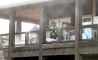 A firefighter wearing gear stands on a wooden balcony with a wire railing. The area is filled with smoke or dust, and various items are stacked against the walls under the roof.