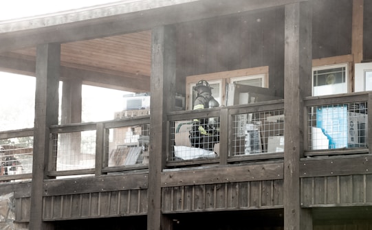 A firefighter wearing gear stands on a wooden balcony with a wire railing. The area is filled with smoke or dust, and various items are stacked against the walls under the roof.